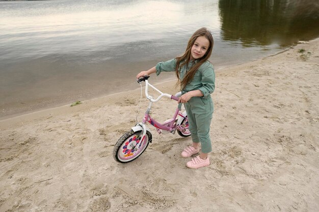 Niña alegre feliz con su bicicleta en la banda del río en la naturaleza