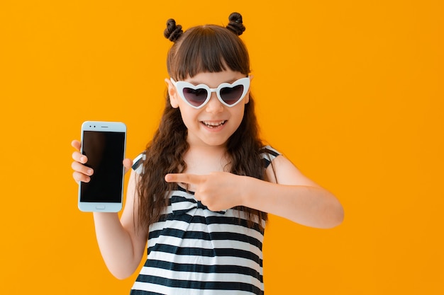 Niña alegre en gafas de sol con un corazón en una pared amarilla señala con el dedo la pantalla del teléfono inteligente. El concepto de servicios y aplicaciones móviles. verano, internet, web