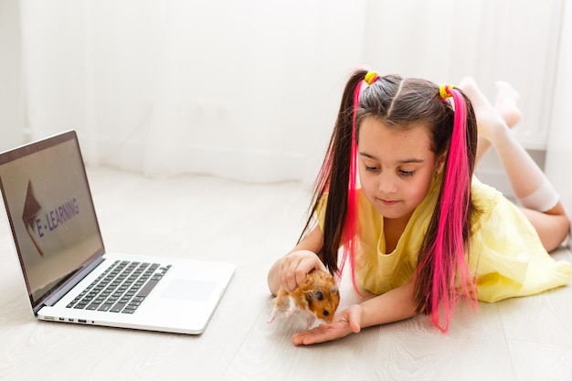 Niña alegre con un hámster mascota usando computadora portátil estudiando a través del sistema de e-learning en línea en casa. Aprendizaje a distancia o remoto