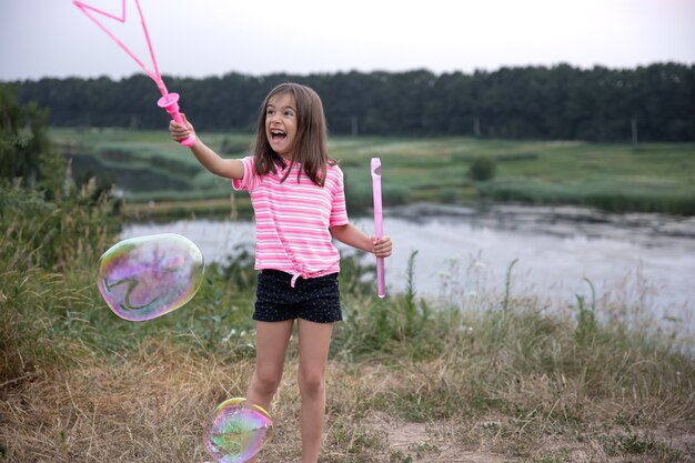 Niña alegre juega con grandes pompas de jabón en la naturaleza.