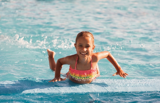 Niña alegre jugando en la piscina con agua clara y clara y mirando sonriendo a la cámara