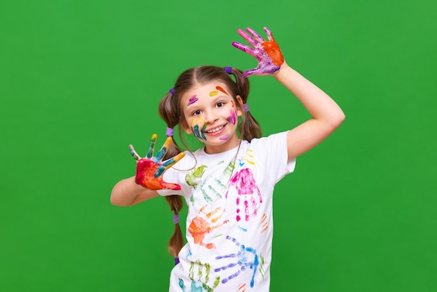 Una niña alegre con las manos manchadas de pintura hace muecas y sonríe una niña se ensució en un estante sobre un fondo verde aislado