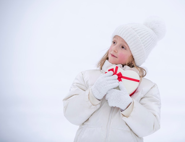 Una niña alegre con ropa blanca de invierno se para frente a la nieve y sostiene una caja en forma de corazón