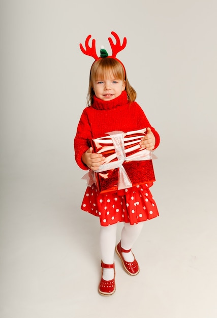 Niña alegre con un suéter rojo sostiene una caja de regalo roja sobre un fondo blanco.