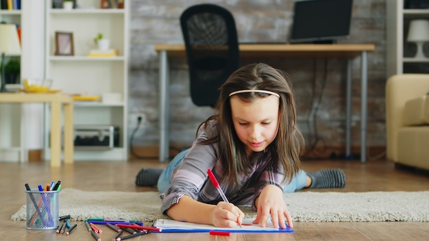 Niña alegre tirada en el suelo haciendo un dibujo creativo.