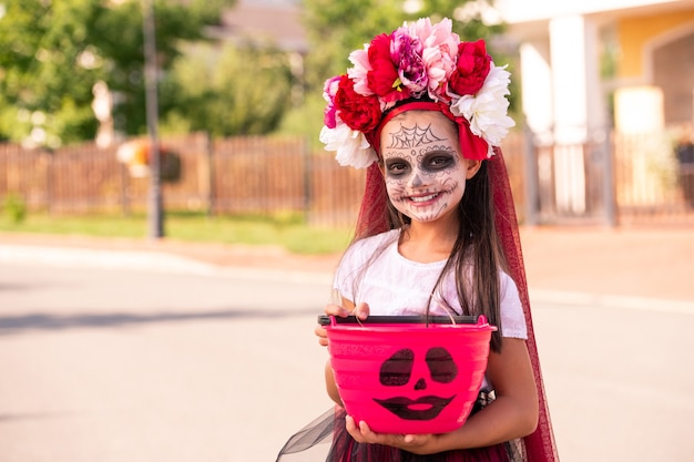 Niña alegre en traje de bruja de halloween sosteniendo un cubo de plástico rosa lleno de dulces mientras está de pie en la carretera contra cabañas