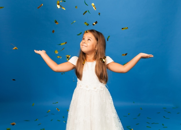 Niña alegre en vestido blanco bailando en confeti aislado sobre fondo azul de estudio.