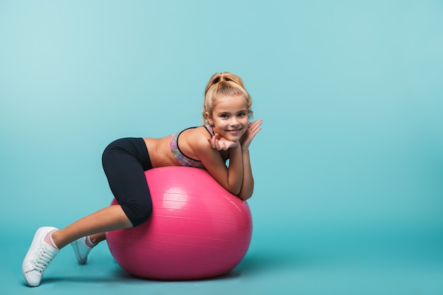 Niña alegre vistiendo ropa deportiva recostada sobre una pelota de fitness aislada sobre pared azul