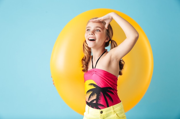 Niña alegre vistiendo traje de baño que se encuentran aisladas sobre la pared azul, jugando con un anillo inflable