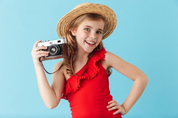 Niña alegre vistiendo traje de baño y sombrero de verano que se encuentran aisladas sobre la pared azul, tomando fotografías con cámara de fotos