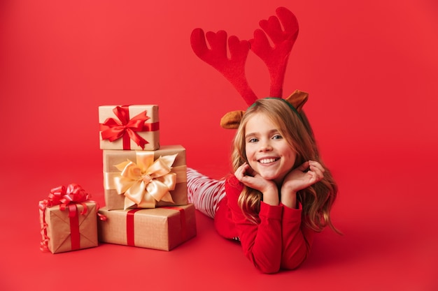 Niña alegre vistiendo traje de ciervo de Navidad sentado con pila de cajas de regalo aislado