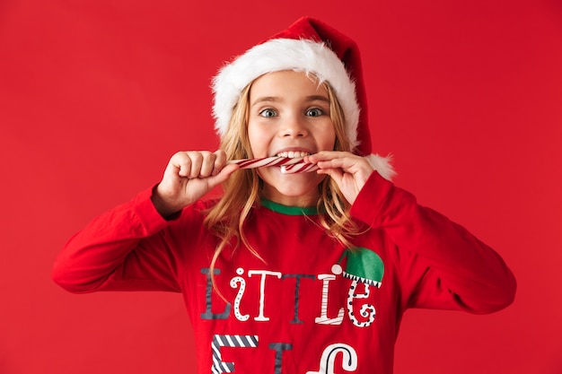 Foto niña alegre vistiendo traje de navidad que se encuentran aisladas, comiendo caramelos de cono de caramelo de navidad