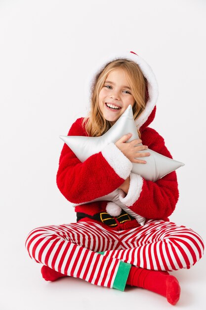 Niña alegre vistiendo traje de Navidad sentado aislado, sosteniendo una almohada en forma de estrella