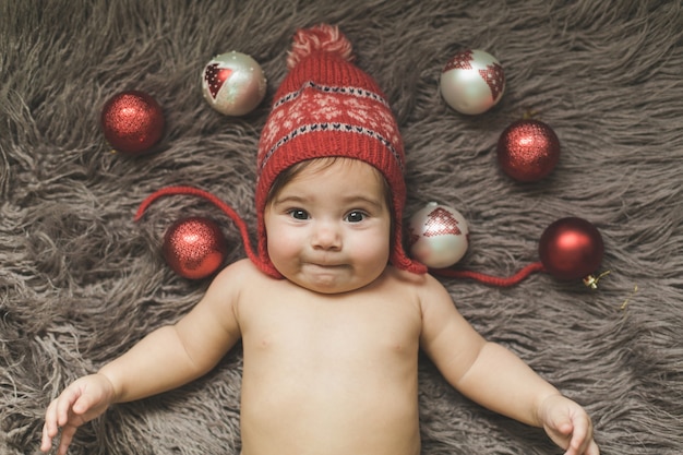 Niña de un año se acuesta en una colcha, con un sombrero rojo y juega con juguetes de Año Nuevo