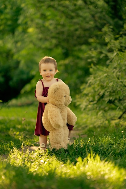 Una niña de un año da sus primeros pasos independientes en un parque soleado agarrando a su oso de peluche frien