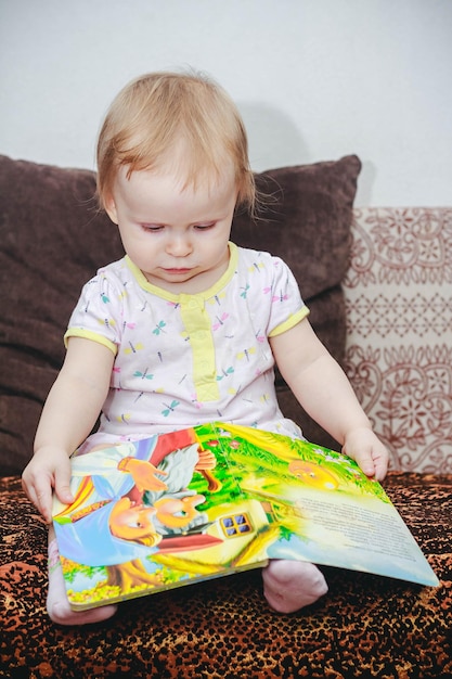 Una niña de un año está sentada en el sofá mirando un libro ilustrado