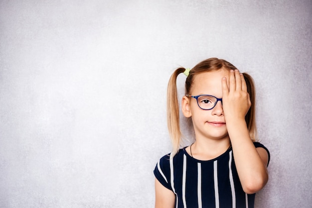 Niña en anteojos cubriendo su ojo con la mano mientras toma el examen de la vista antes de la escuela, el jardín de infantes y el chequeo médico escolar