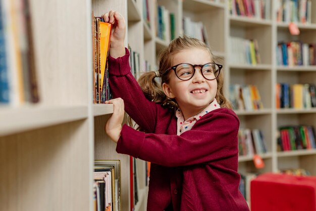 Niña con anteojos toma un libro de una estantería en una biblioteca o aula