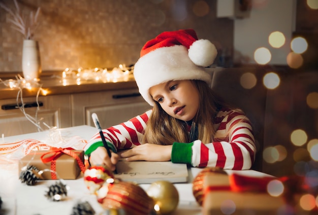 Una niña antes de Navidad escribe una carta a Santa Claus sobre la mesa con guirnaldas. niño escribe una carta a santa claus.