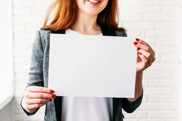 Una niña de apariencia europea sostiene una hoja de papel en blanco para la colocación de una inscripción, publicidad, lema, marca registrada