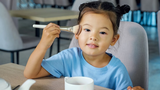 La niña aplica maquillaje. Lindo niño asiático con bollos espaciales mueve un cepillo grande con polvo en las mejillas y mira recto sentado en la mesa de café vista cercana