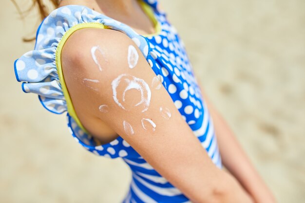 Niña con la aplicación de protector solar en el hombro en forma de sol de pie en la playa. Vacaciones de verano.