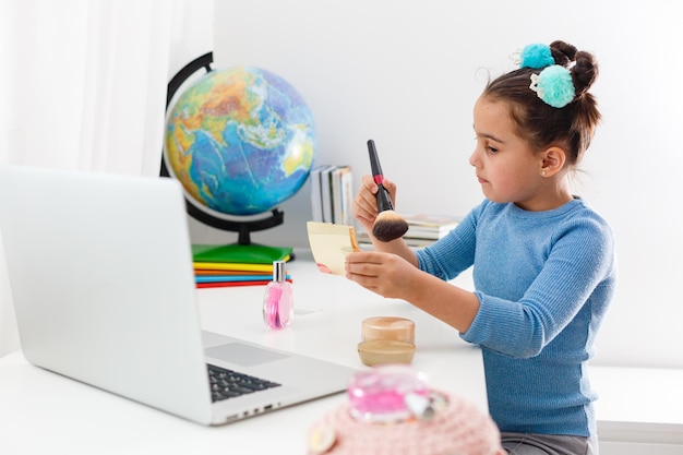 niña aprendiendo maquillaje en polvo, brocha, rubor en computadora portátil en línea, aprendizaje a distancia