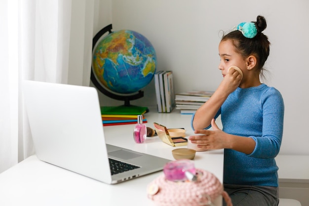 niña aprendiendo maquillaje en polvo, brocha, rubor en computadora portátil en línea, aprendizaje a distancia