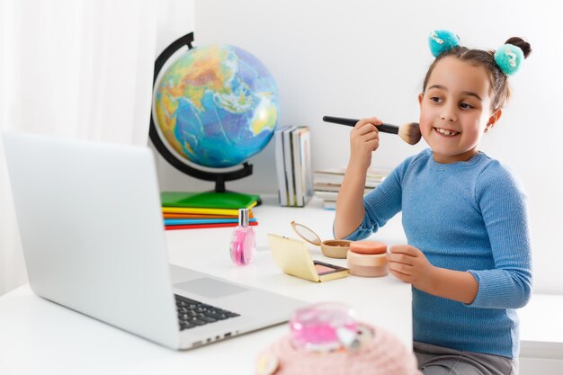 niña aprendiendo maquillaje en polvo, brocha, rubor en computadora portátil en línea, aprendizaje a distancia