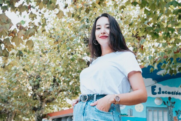 Niña árabe africana entre hojas verdes sonriendo. Señalando Copiar espacio para texto y agrega. Ropa de moda, jeans y camiseta blanca de estilo moderno. Redes sociales