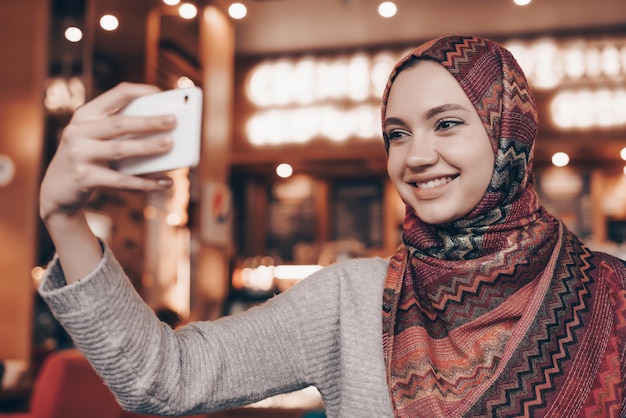 Niña árabe sonriente con un pañuelo en la cabeza sentada en un restaurante acogedor y haciendo selfie en su teléfono inteligente