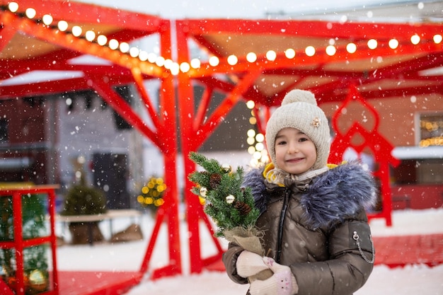 Una niña con un árbol de Navidad en sus manos al aire libre con ropa abrigada en invierno en un mercado festivo Guirnaldas de luces de hadas decoradas ciudad de nieve para el nuevo año