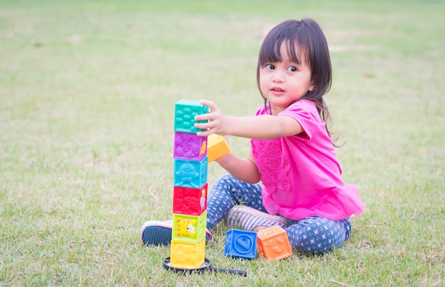 La niña asiática adorable está jugando los bloques coloridos en el campo de hierba en el parque