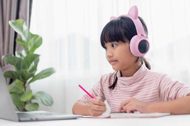 Una niña asiática con auriculares tiene una clase distante de videollamadas con un maestro que usa un estudio portátil en línea sobre el concepto de educación en el hogar por computadora