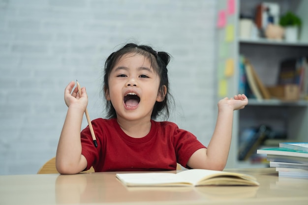 Una niña asiática con una camisa roja escribe notas en un cuaderno y lee un libro para estudiar en línea en un escritorio de madera en la sala de estar en casa Educación aprendiendo en línea desde el concepto de hogar