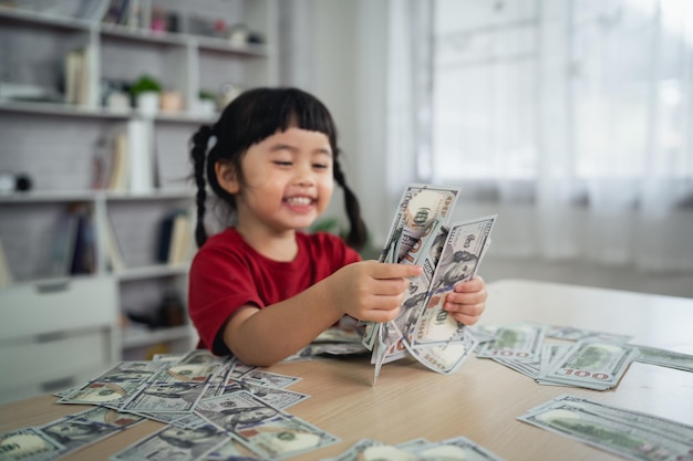 Niña asiática con una camiseta roja sosteniendo un billete de un dólar en un escritorio de madera en la sala de estar en casa