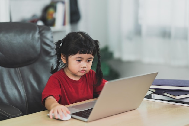 Una niña asiática con una camiseta roja usa una computadora portátil y estudia en línea en un escritorio de madera en la sala de estar en casa Educación aprendiendo en línea desde el concepto de hogar