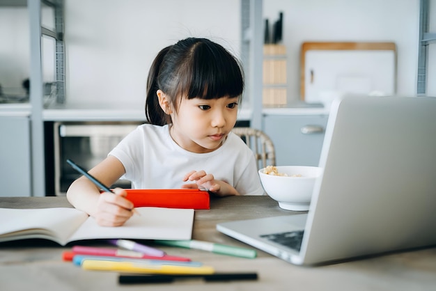 Una niña asiática desayunando mientras estudia desde casa Asiste a clases escolares en línea con portátil y tableta digital y escribe notas en casa