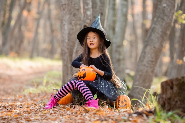 Una niña asiática en un disfraz de bruja para Halloween camina en el bosque con un balde para dulces