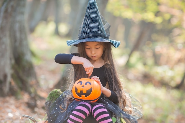 Una niña asiática en un disfraz de bruja para Halloween camina en el bosque con un balde para dulces