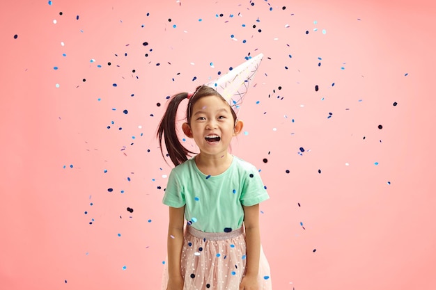 Niña asiática divirtiéndose lanzando confeti con sombrero de fiesta de pie contra el rosa aislado