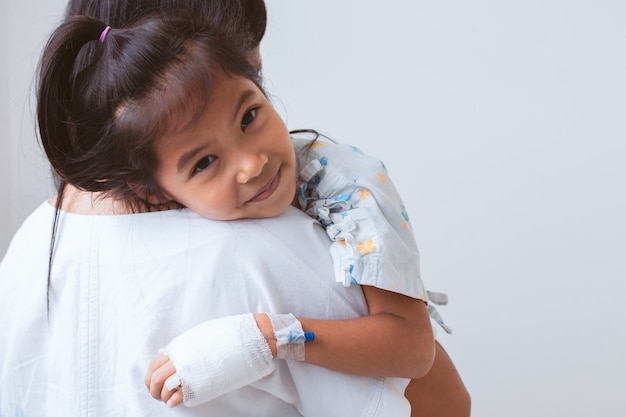 Niña asiática enferma que tiene una solución intravenosa vendada sonriendo y abrazando a su madre en el hospital