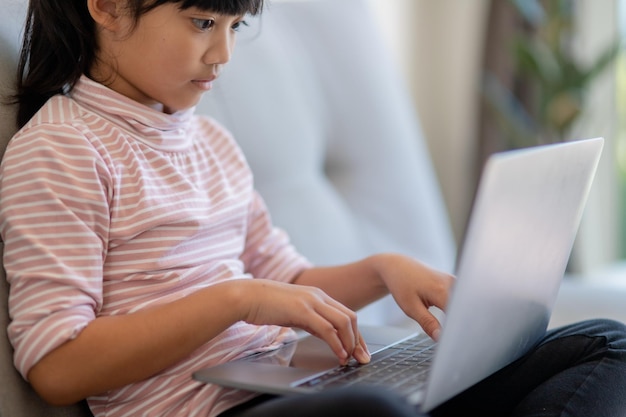 La niña asiática de la escuela pequeña usa una computadora portátil sentada en el sofá sola en casa