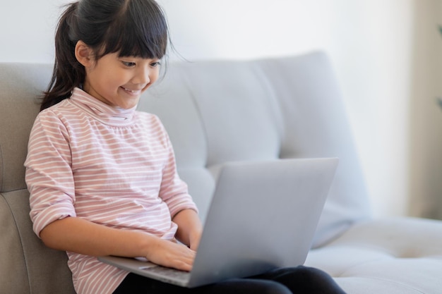 La niña asiática de la escuela pequeña usa una computadora portátil sentada en el sofá sola en casa