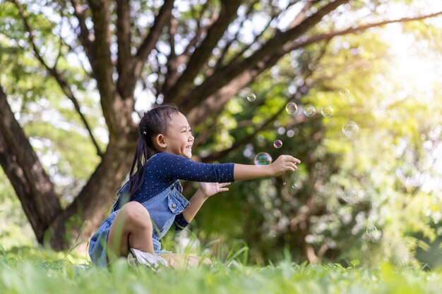 Niña asiática feliz niño soplando pompas de jabón afuera en el parque verde.