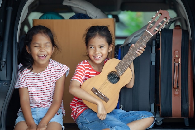 Niña asiática feliz tocando la guitarra y cantando una canción con su hermana en el maletero de un coche
