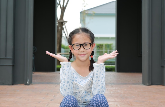 Niña asiática con gafas con las manos abiertas y mirando la cámara mientras se sienta en un pabellón en el jardín al aire libre