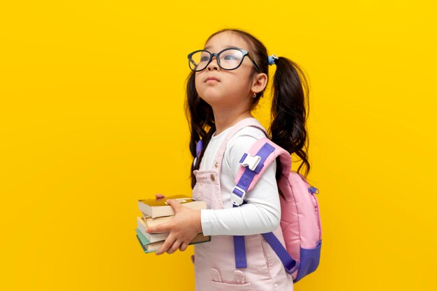 Una niña asiática con gafas y mochila sosteniendo libros sobre un fondo amarillo aislado