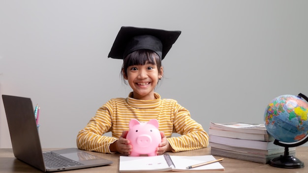 Niña asiática con una gorra de graduación con una alcancía rosa Ahorrando dinero invirtiendo en el futuro