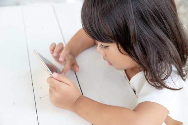 Niña asiática jugando con el teléfono inteligente en la mesa viendo el teléfono inteligente Los niños usan teléfonos y juegan juegos